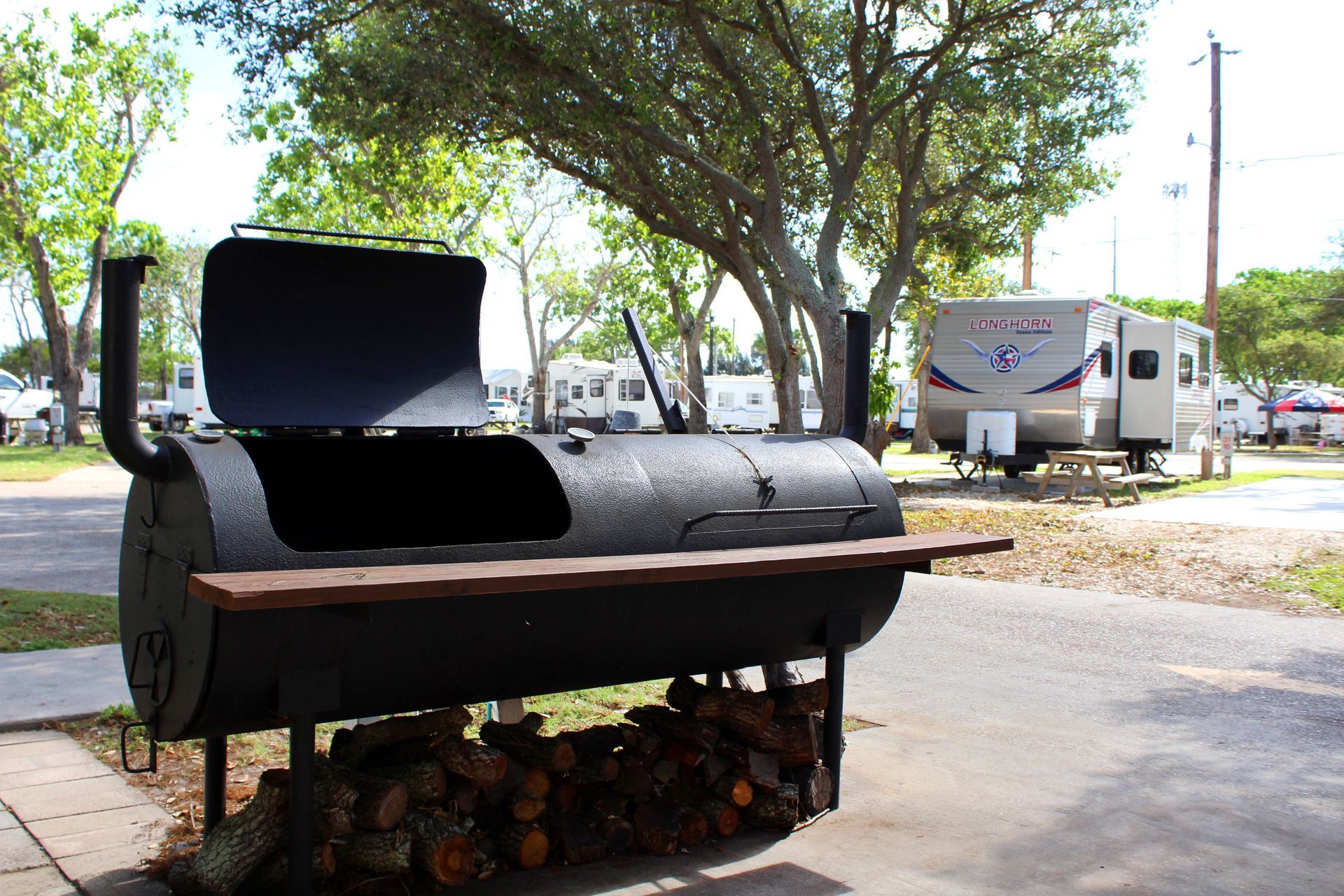 Black barrel smoker grill outdoors under trees, with RVs in the background.
