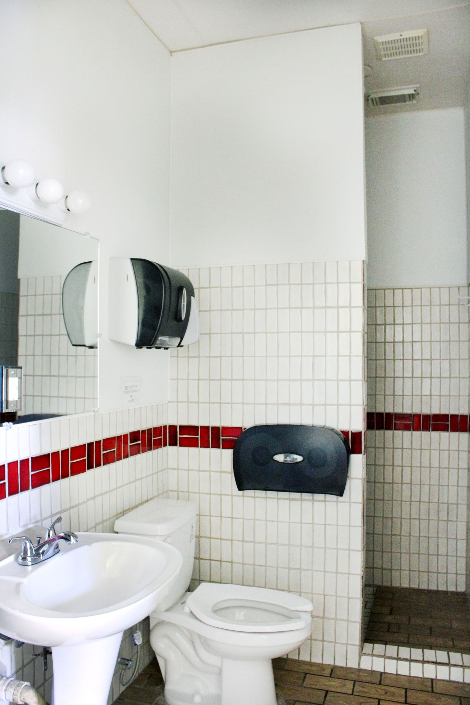 Small public restroom with white tiled walls, red accent band, sink, toilet, mirror, paper towel dispenser, and shower stall