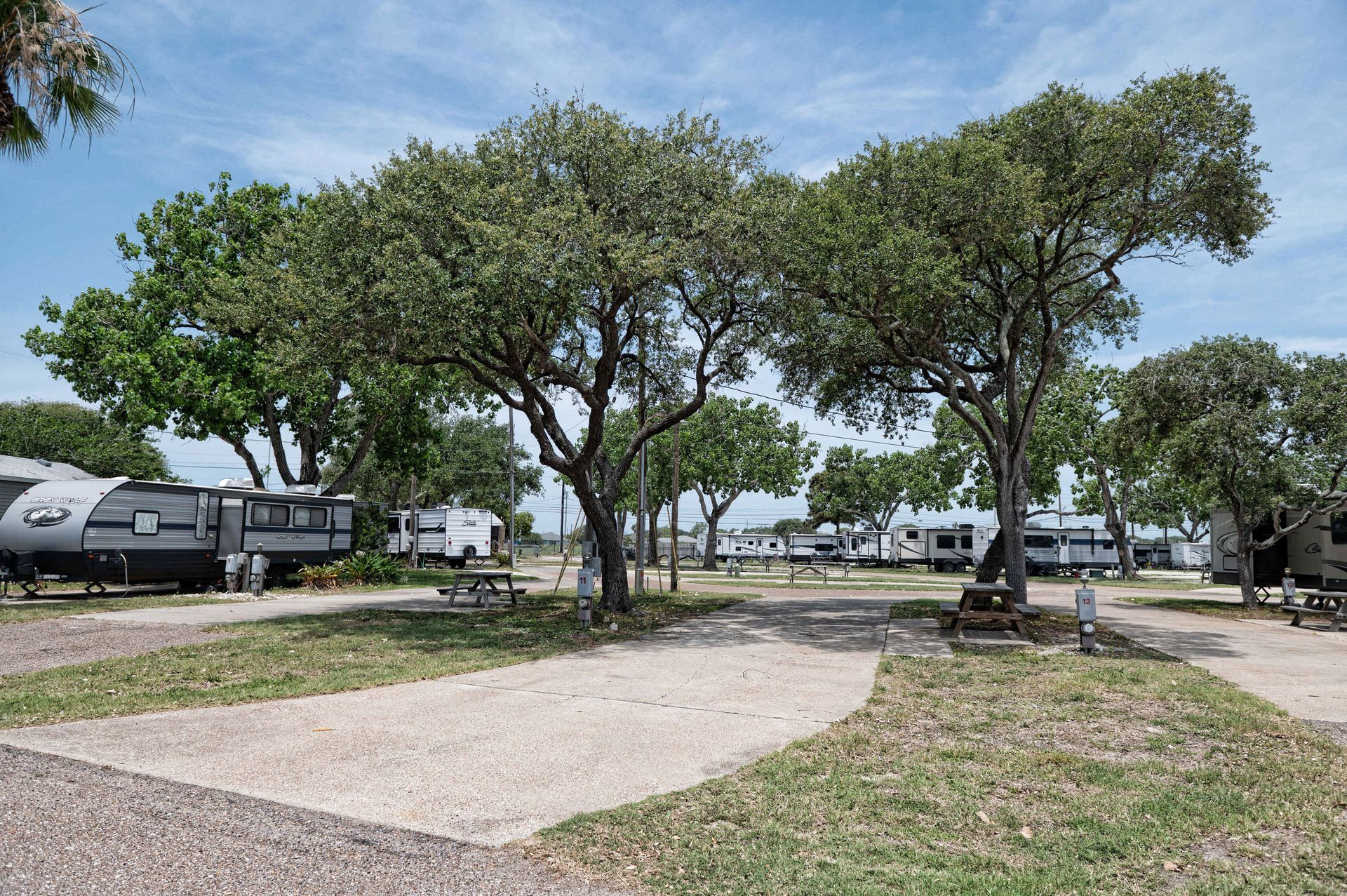 Shaded campground with trees, RVs, gravel paths, and picnic tables under a blue sky