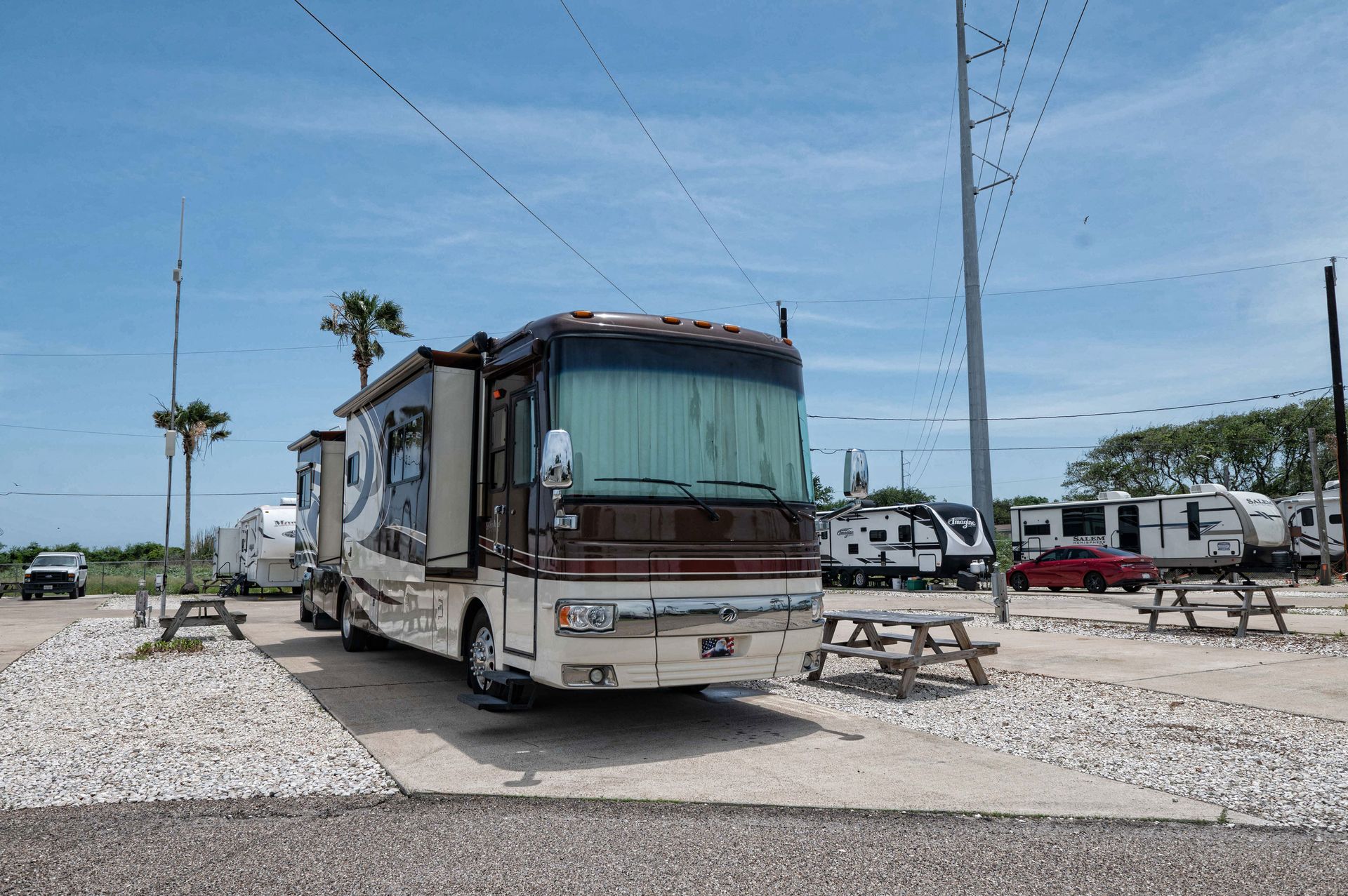 White RV parked on gravel at a sunny campground with palm trees and a picnic table nearby