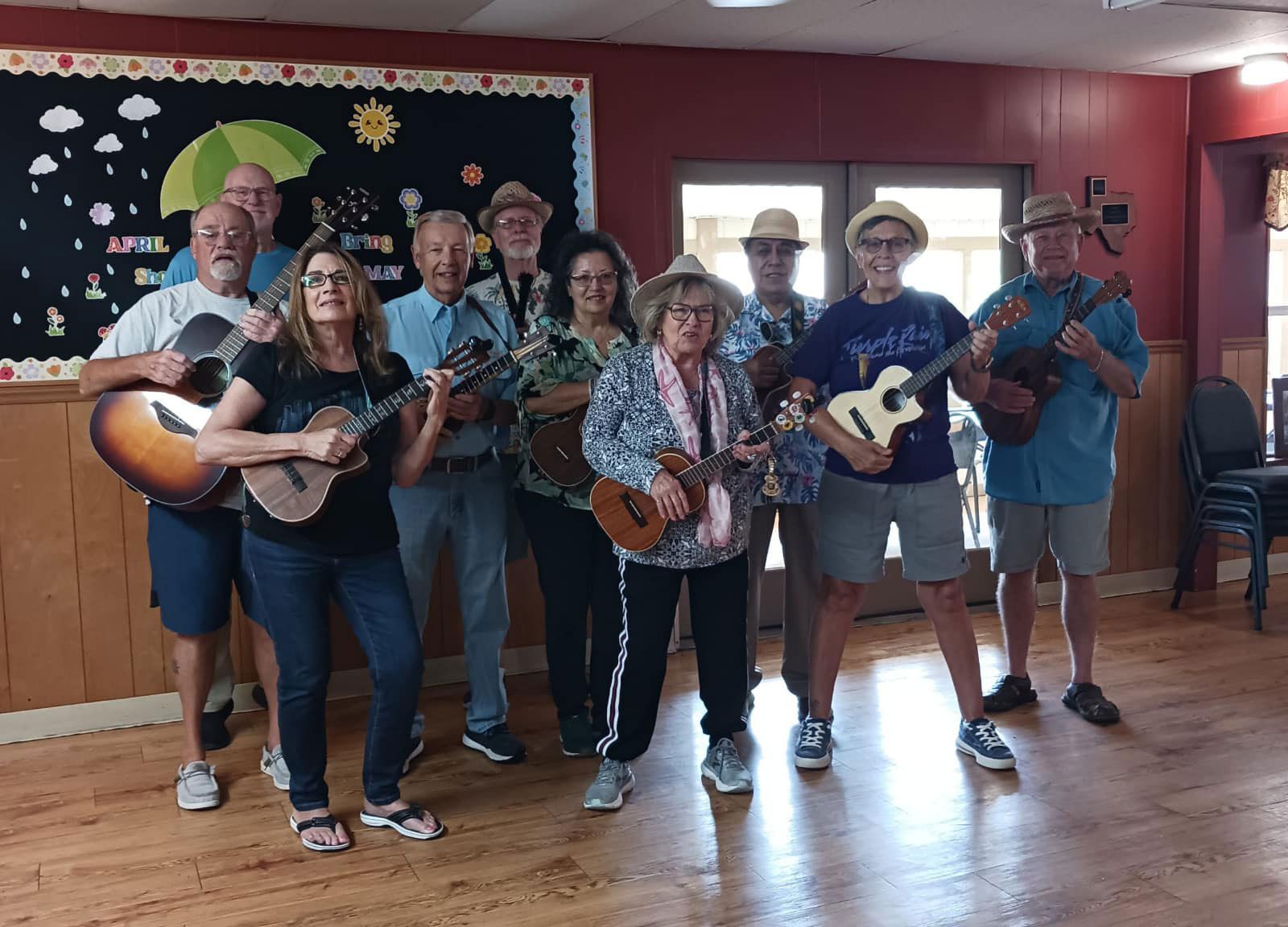 Seven people holding guitars and a ukulele pose in a room with wooden floors and a red wall.