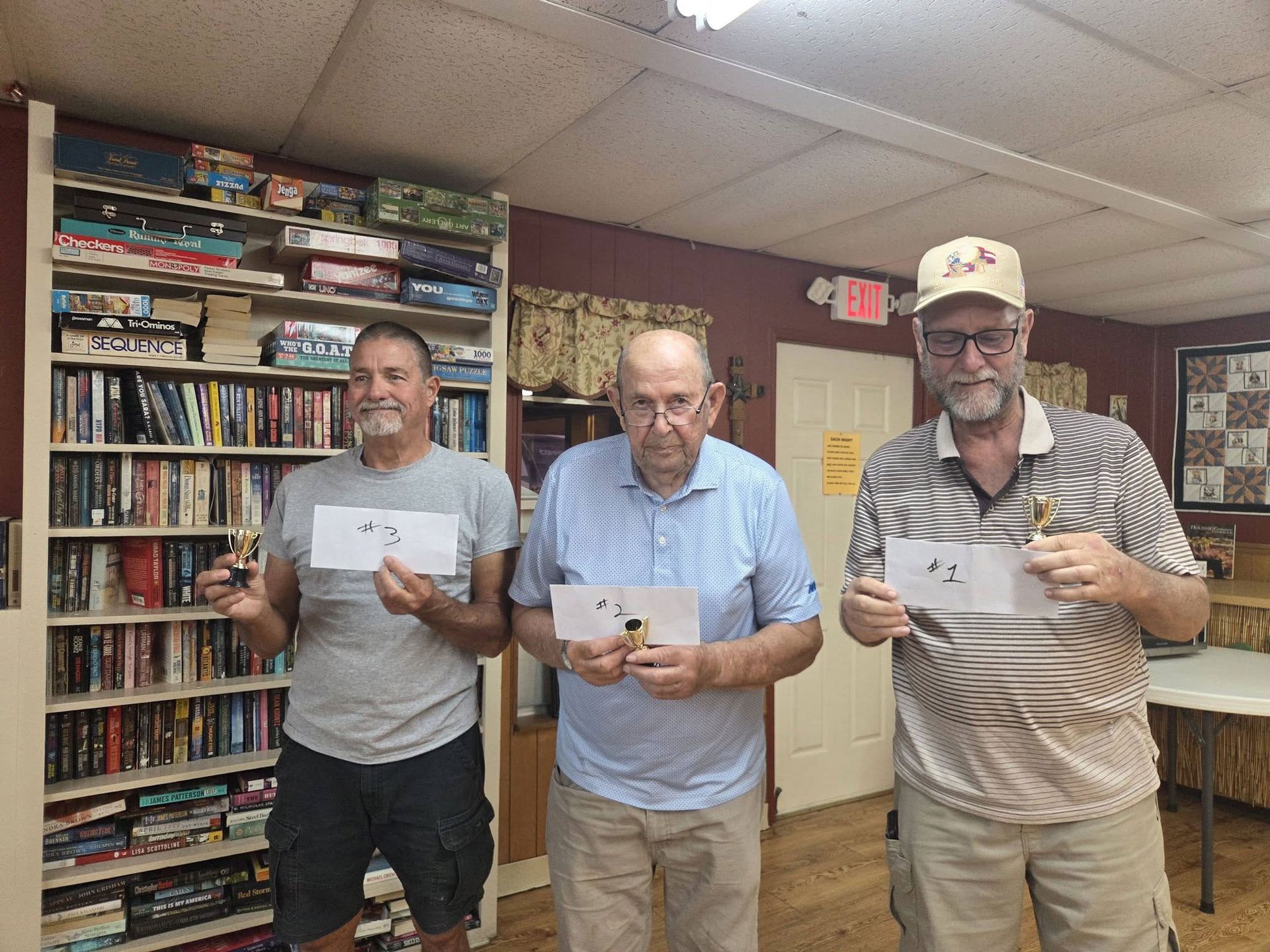 Three men standing indoors in a room with shelves, each holding a small card and facing the camera.