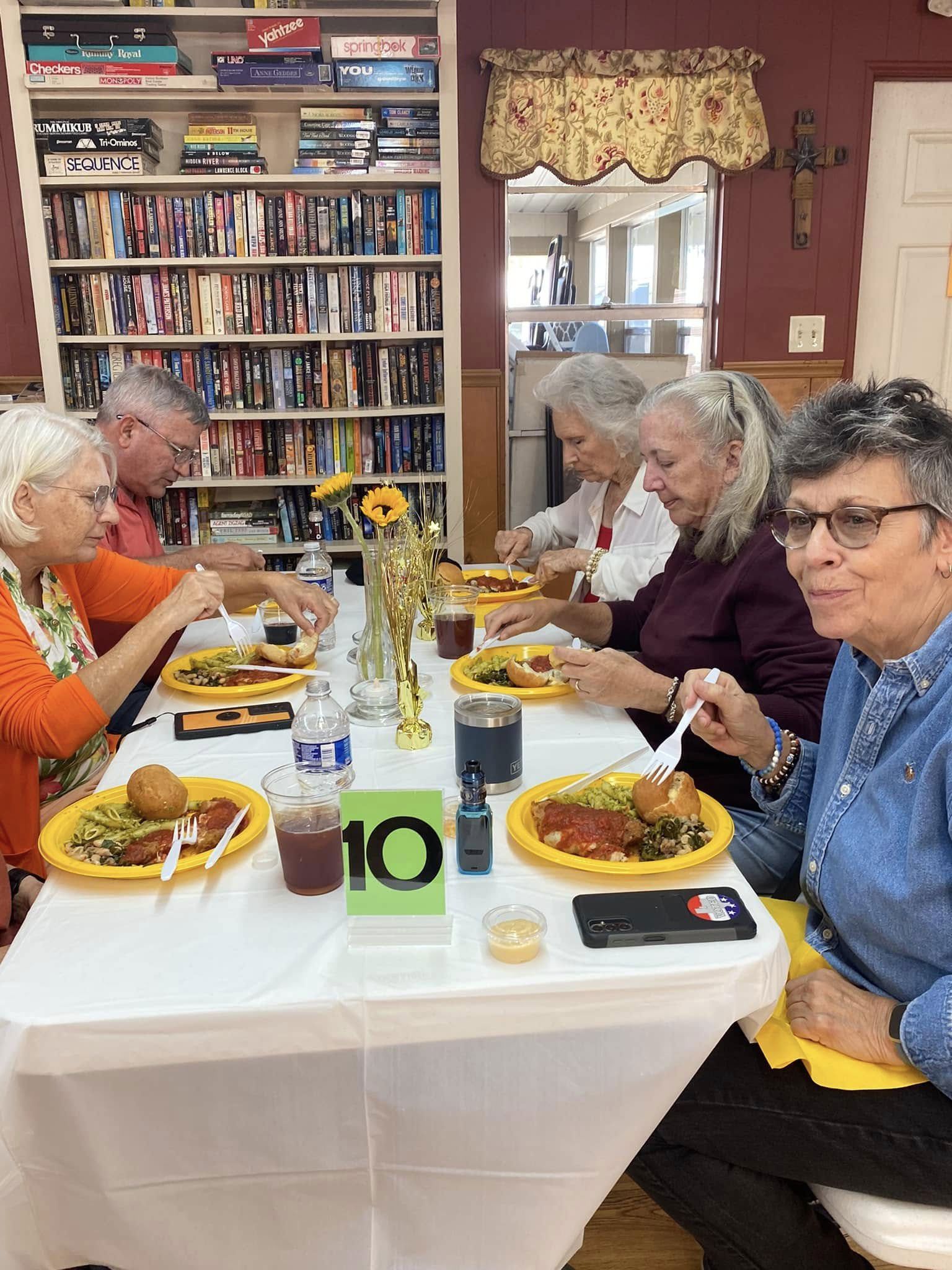 People dining around a table in a cozy room with bookshelves and flowers, plates served with food.