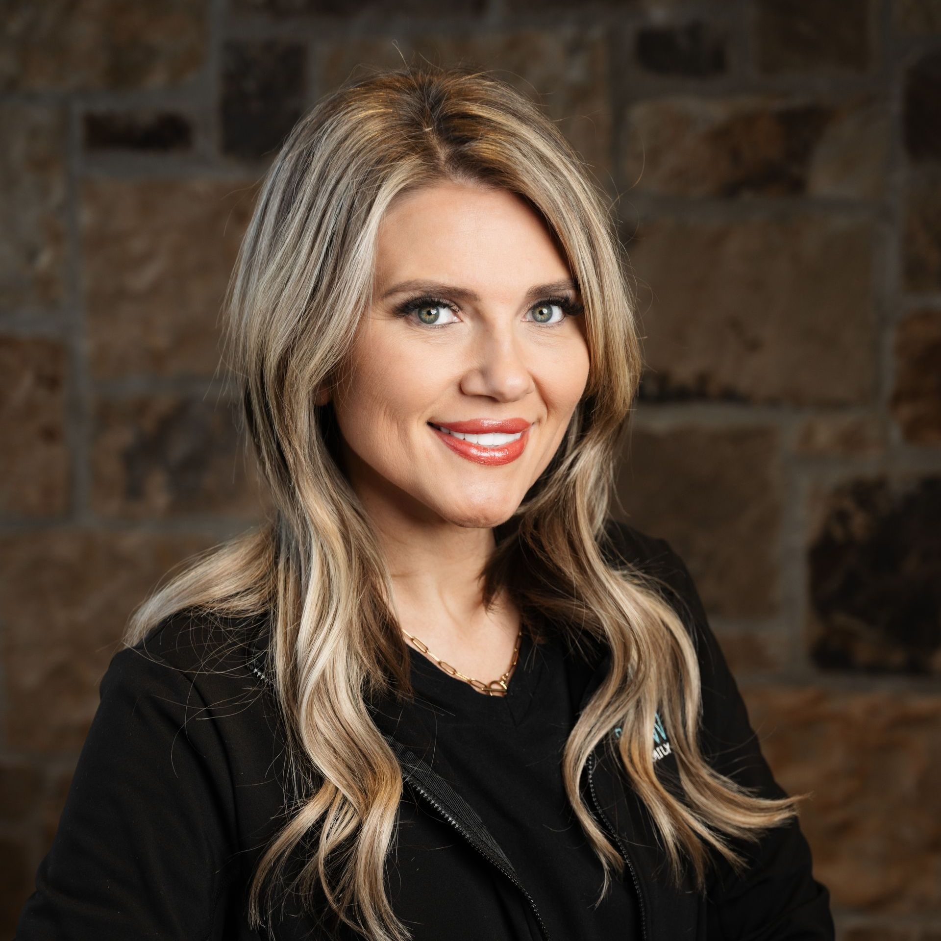 Blonde woman with fair skin smiles, wearing black, in front of a stone wall.