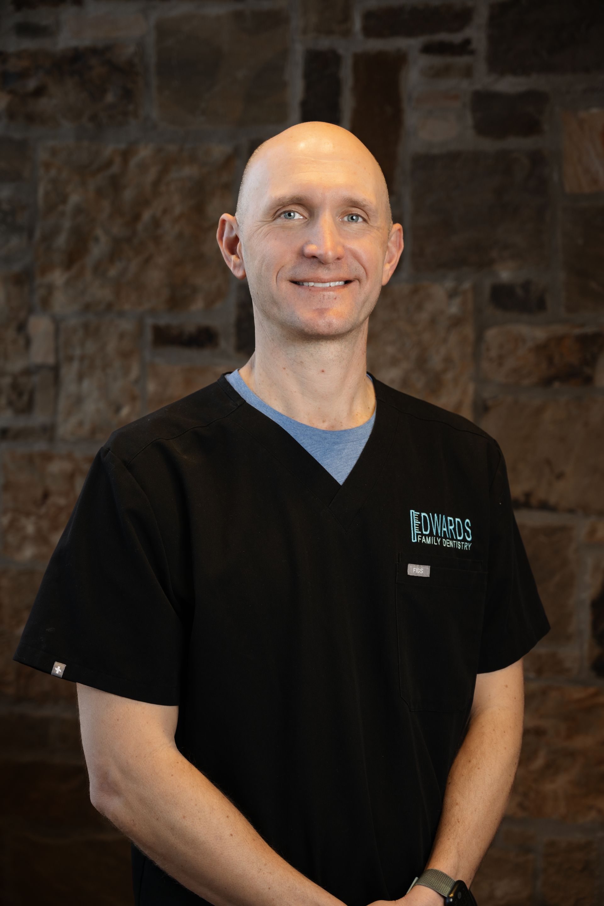 Bald man in black scrubs, smiling, arms at sides, standing in front of a stone wall.