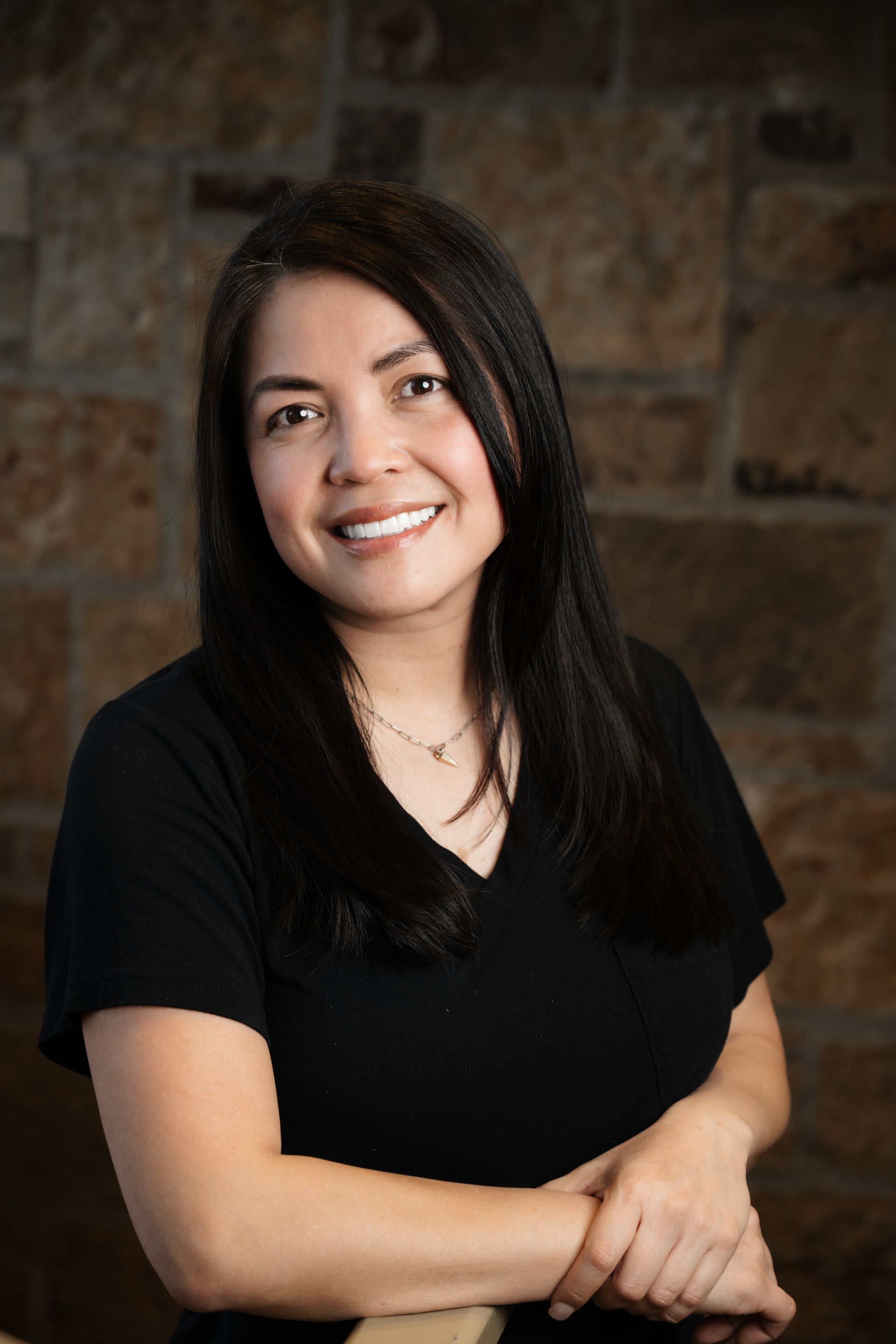 Woman with long dark hair smiles, wearing a black shirt, posing in front of a stone wall.