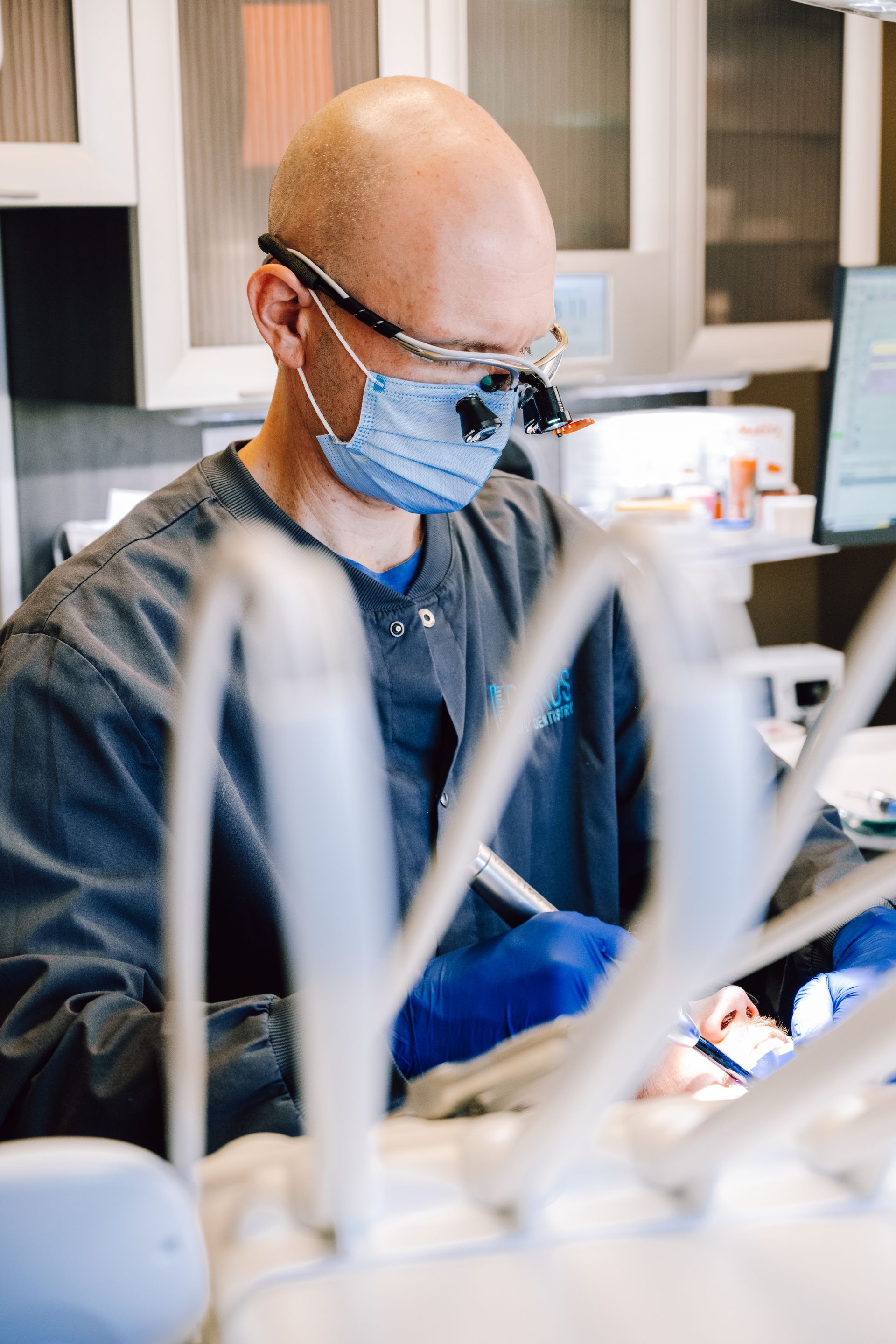 Dentist in scrubs, mask, and loupes, working on a patient's teeth in a dental office.