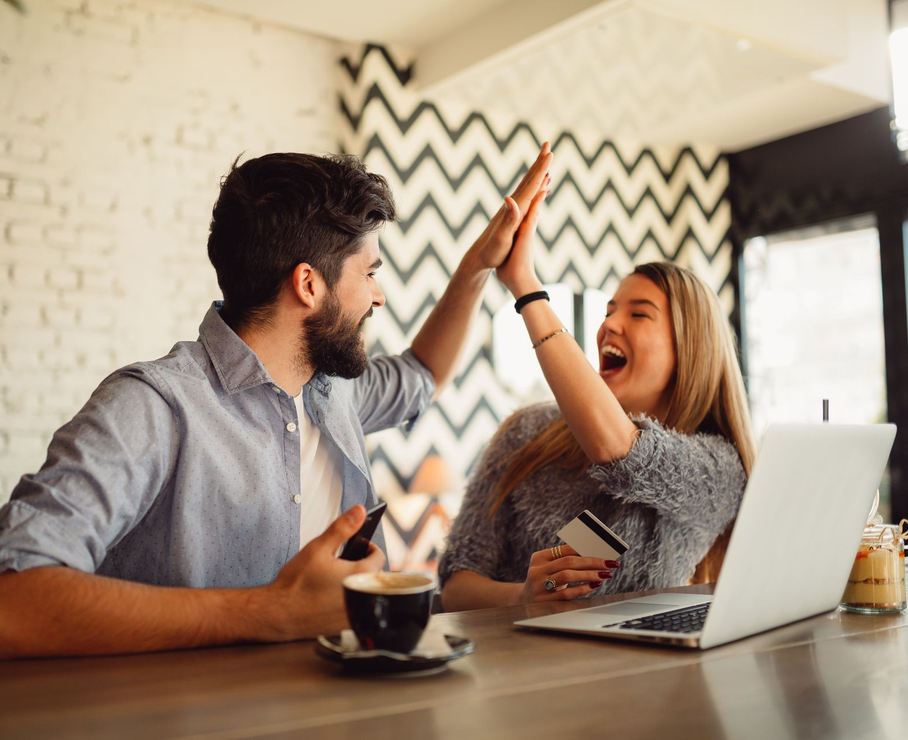 Two coworkers high-five at a desk with a laptop and coffee in a bright office
