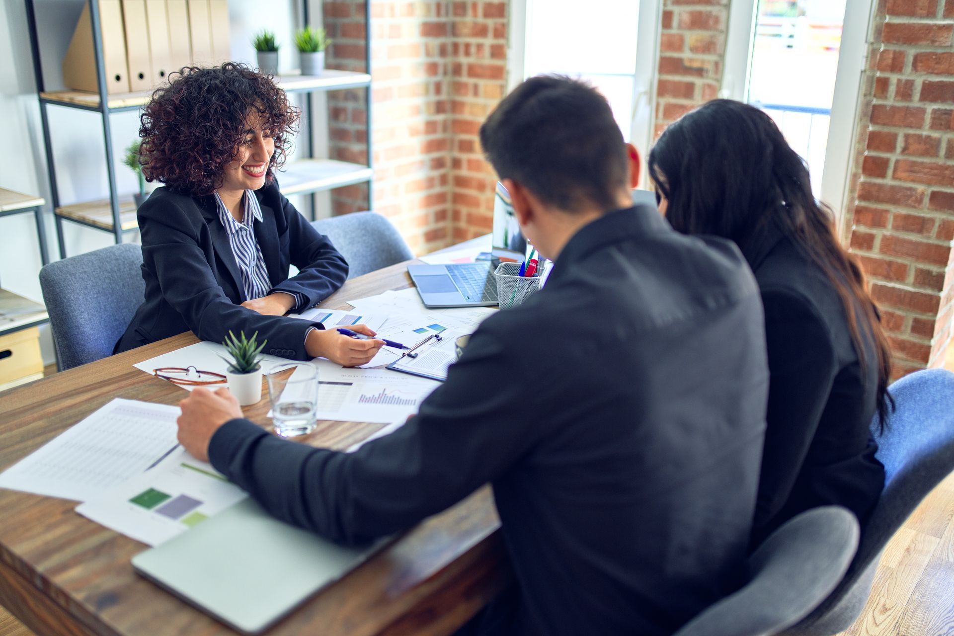 Three people in a bright office meeting around a wooden table with papers and a laptop.