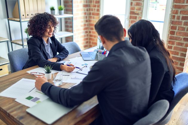 Three people in a bright office meeting around a wooden table with papers and a laptop.