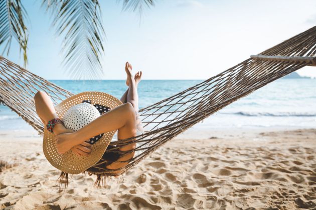 Person relaxing in a beach hammock under palm fronds, feet raised, with ocean and sand in the background