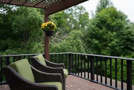 Deck with green cushioned chairs, overlooking lush green trees, yellow hanging flowers.
