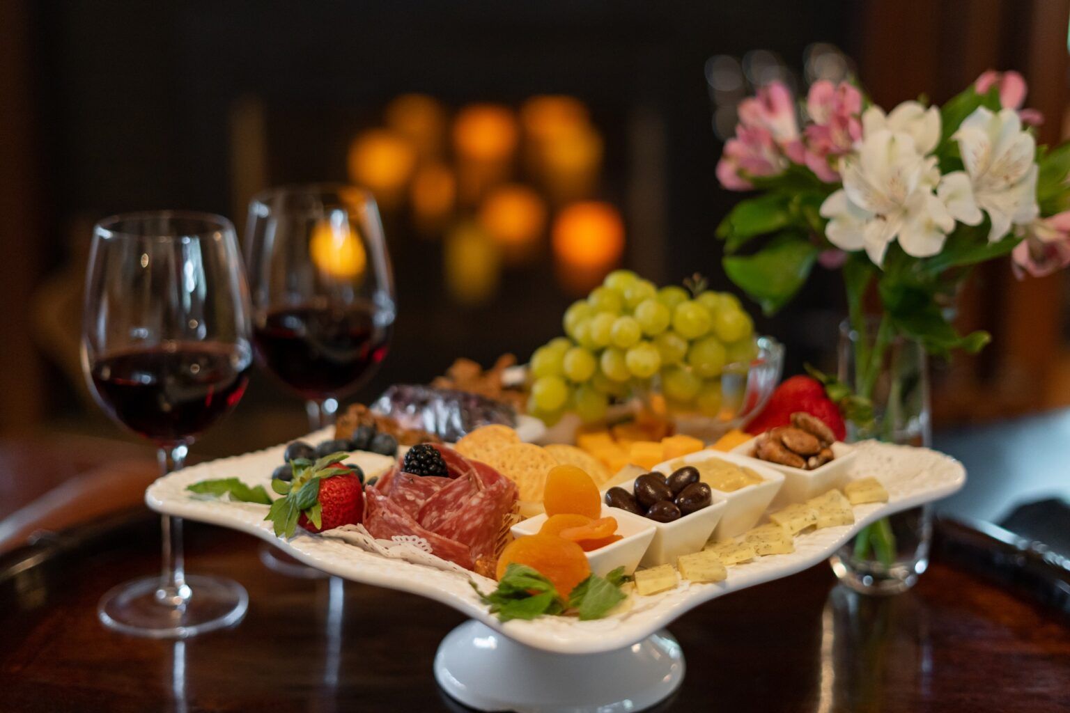 A charcuterie plate with cheese, meats, and fruits, and wine glasses on a table in front of a fireplace.