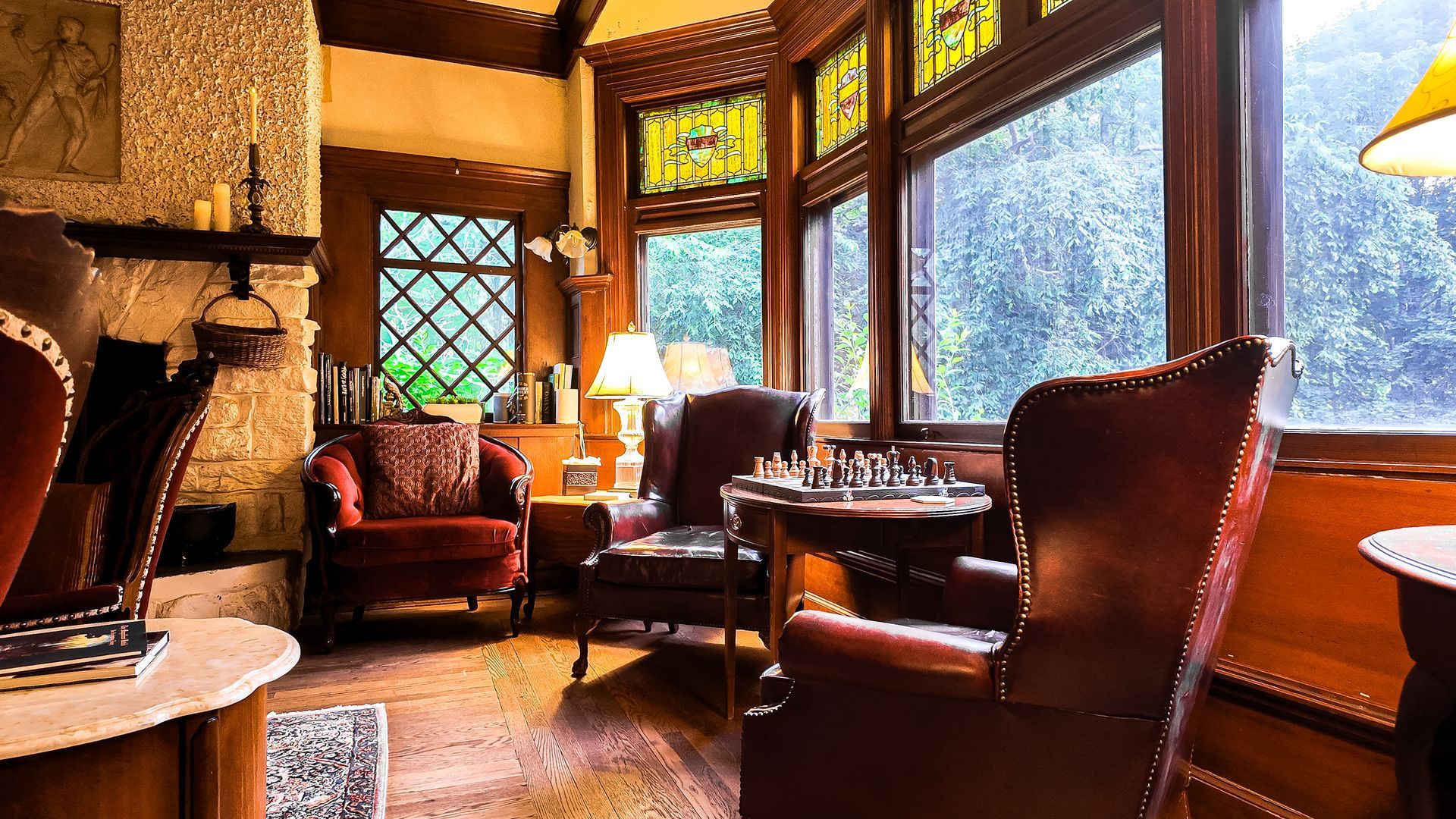 Living room with leather chairs, chess set, fireplace, and stained-glass windows overlooking a wooded area.