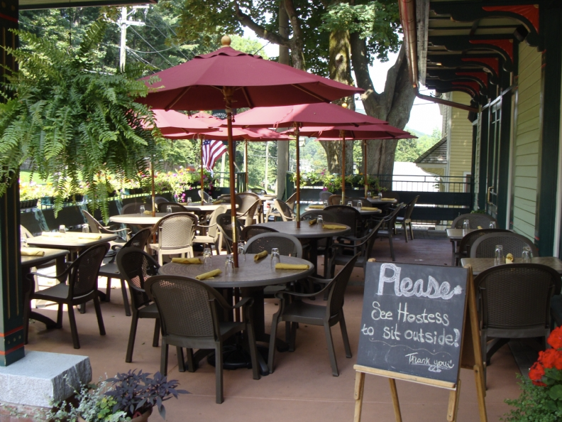 Outdoor café patio with tables, chairs, red umbrellas, and a chalkboard menu sign