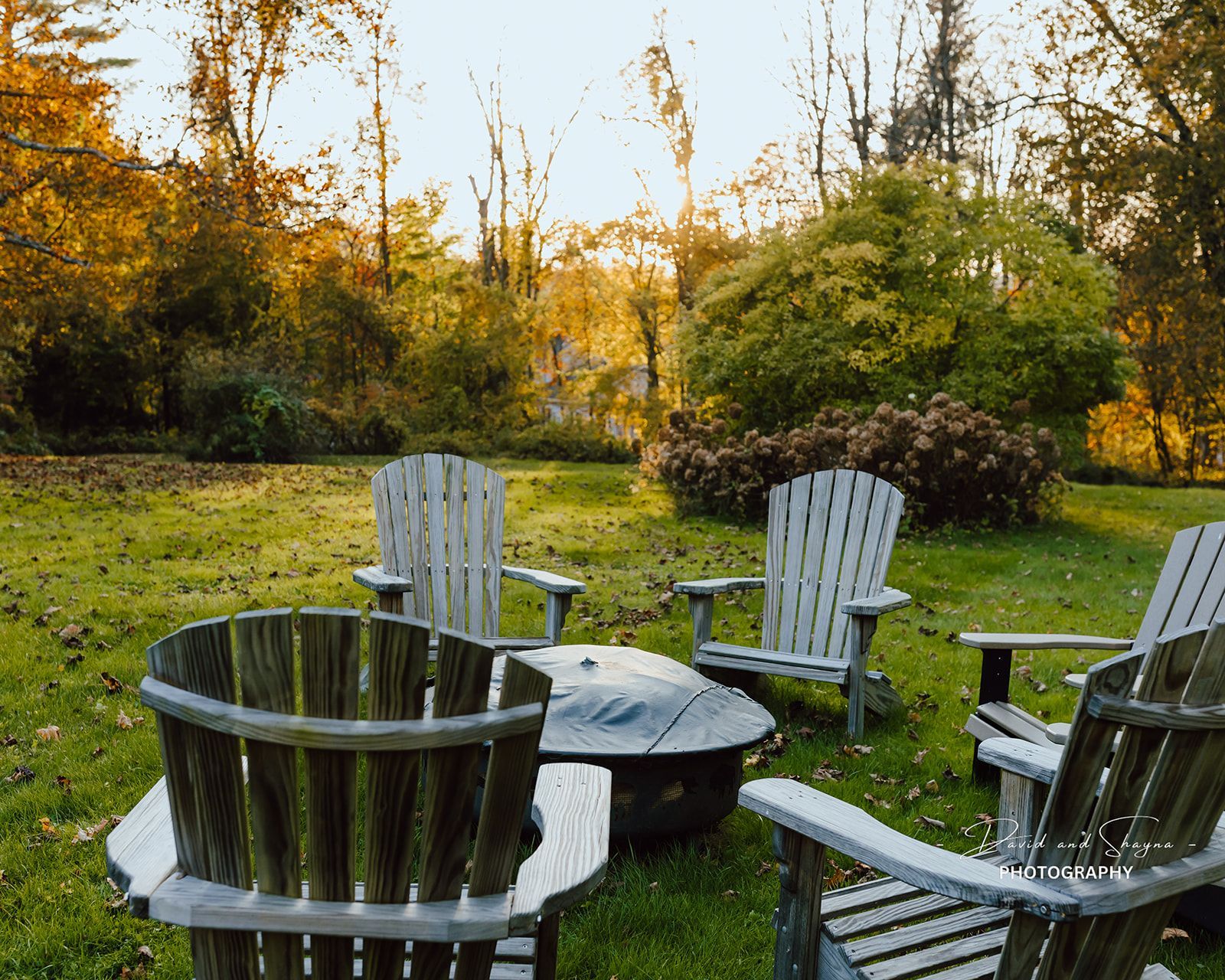 Four wooden Adirondack chairs circle a fire pit on a grassy lawn, autumn foliage in the background.