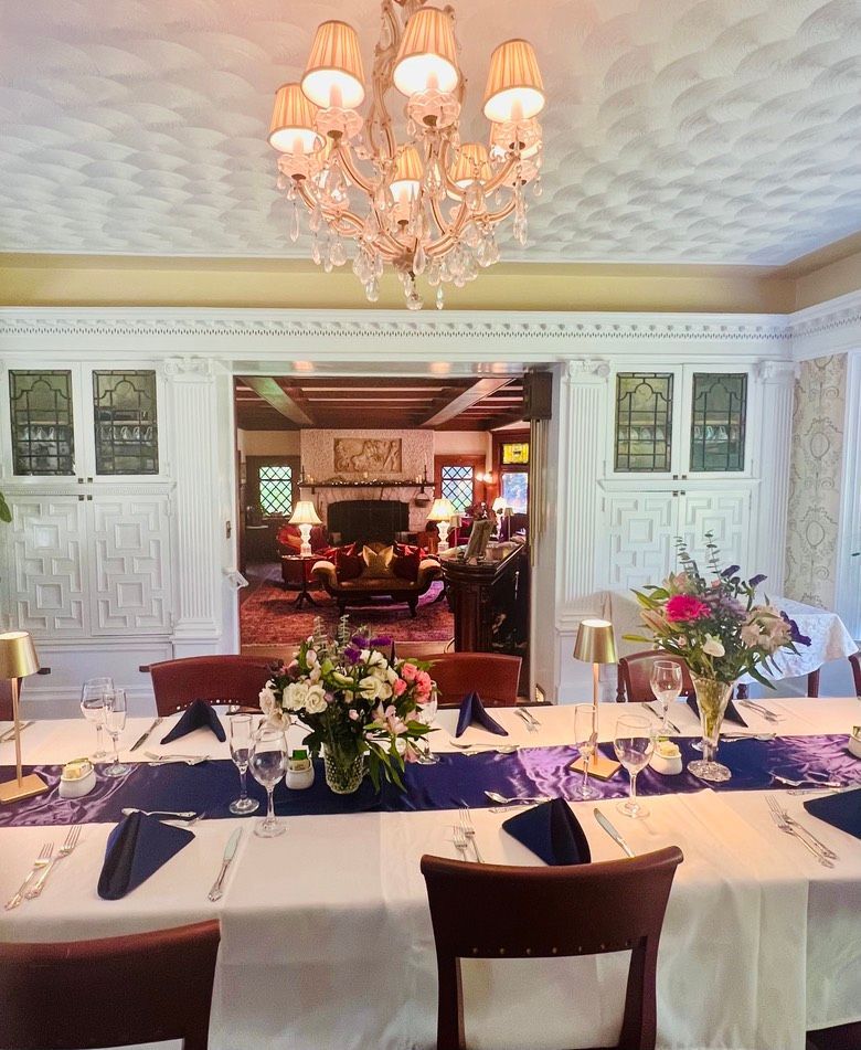 A formal dining room with a long table set for a meal, flowers, and a view into a living room with a fireplace.