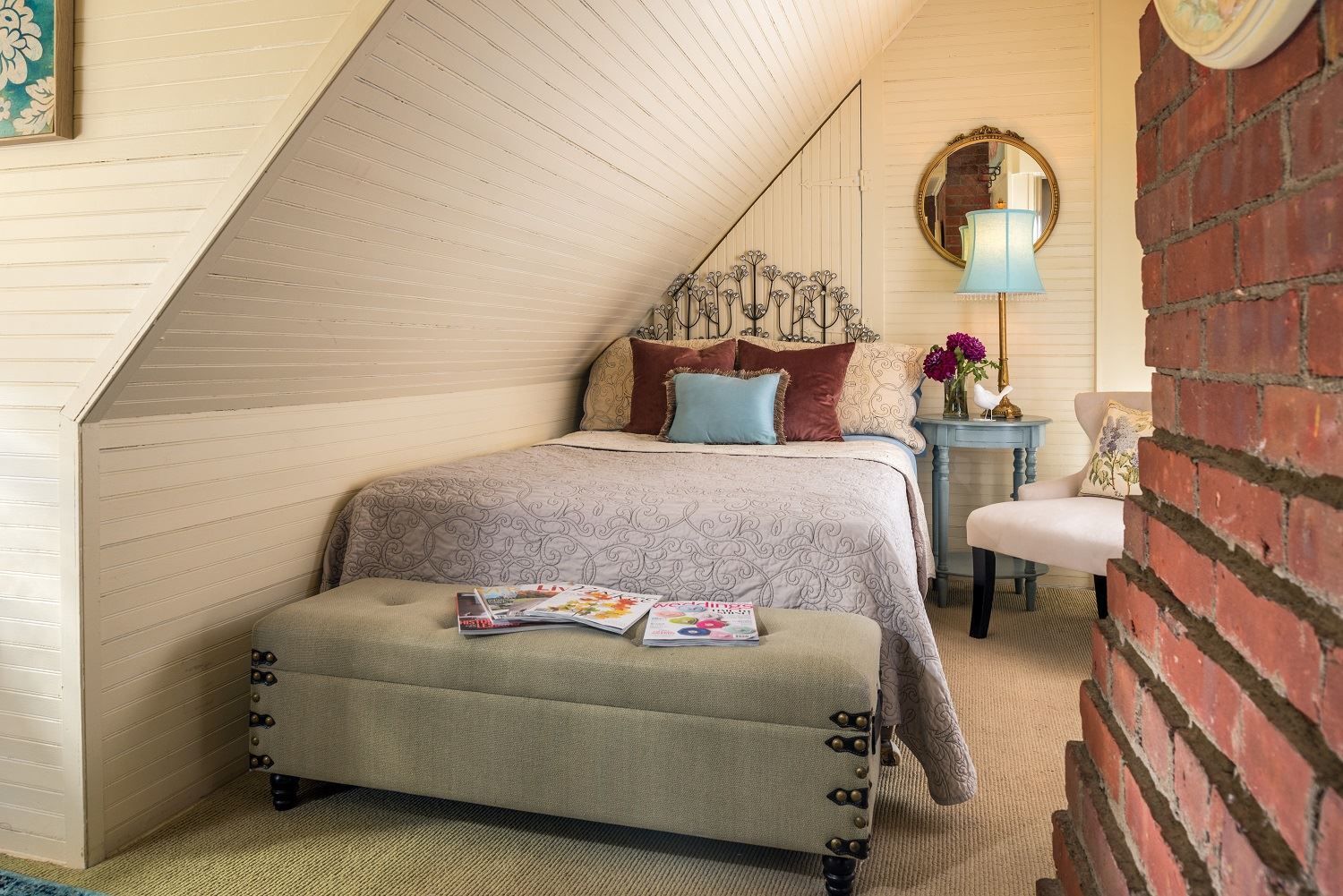 A bedroom at a New England Country Inn with a bed, ottoman, chair and brick wall.