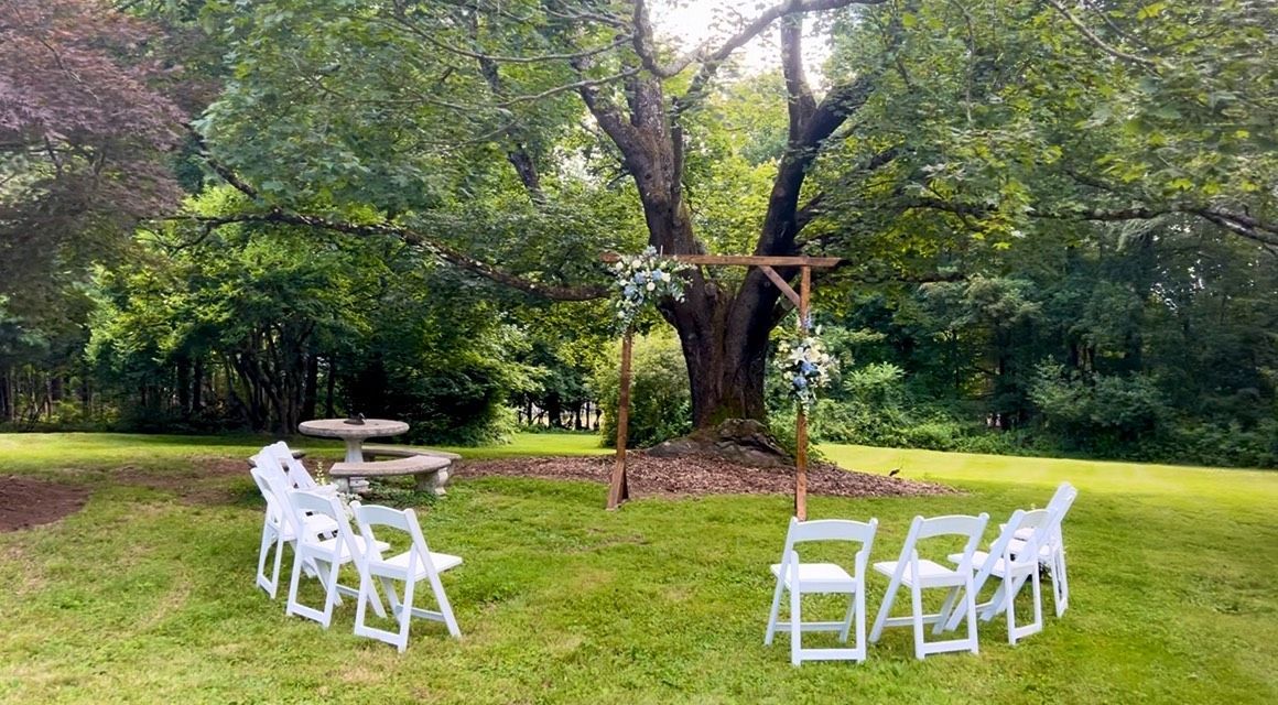 Outdoor wedding setup: white chairs arranged in a circle around a wooden arch decorated with greenery beneath a large tree.
