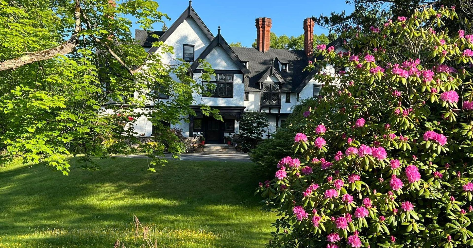 A large white Tudor-style mansion is a New England Country Inn surrounded by trees and flowers.