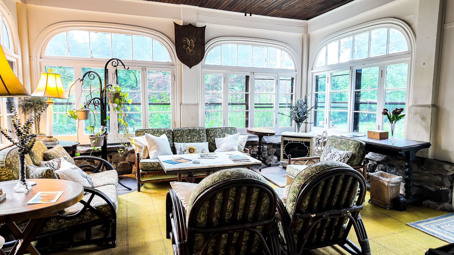 Sunroom with couches, chairs, and tables. Large arched windows overlook greenery. Warm, inviting space.