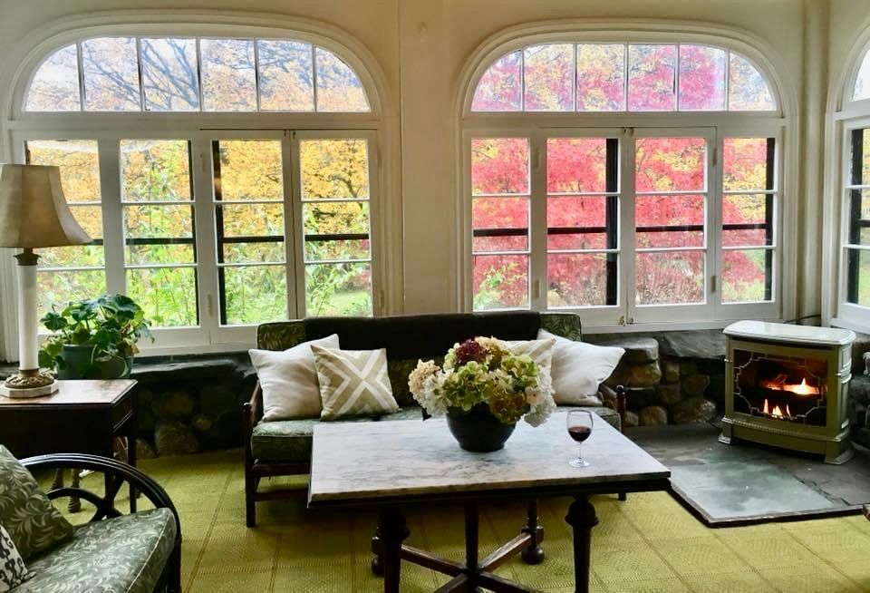 Sunroom with arched windows displaying fall foliage. Green couch, marble table, fireplace.