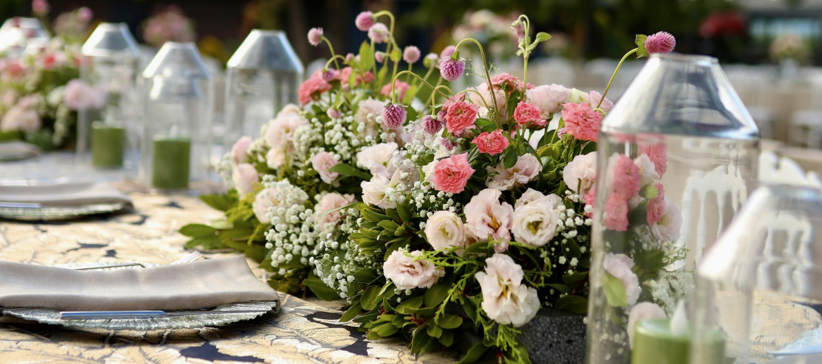 Floral table centerpiece with pink and white flowers beside glass lanterns on a rustic outdoor table