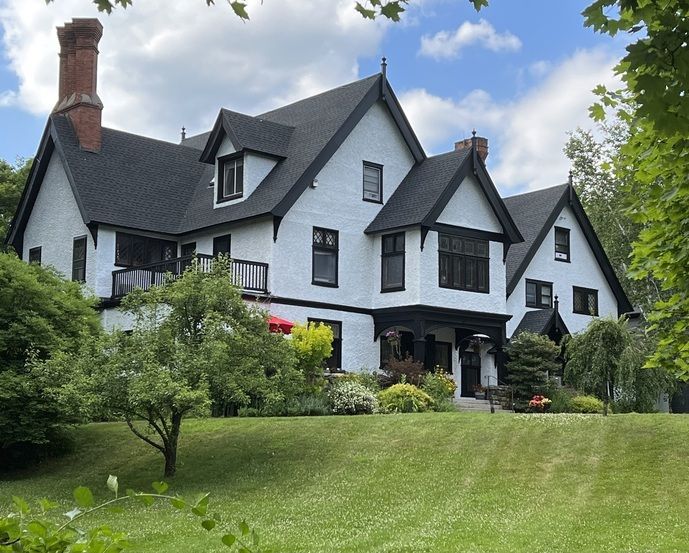 A large white Tudor-style mansion with a black roof is sitting on top of a grassy hill with trees and expansive lawns.