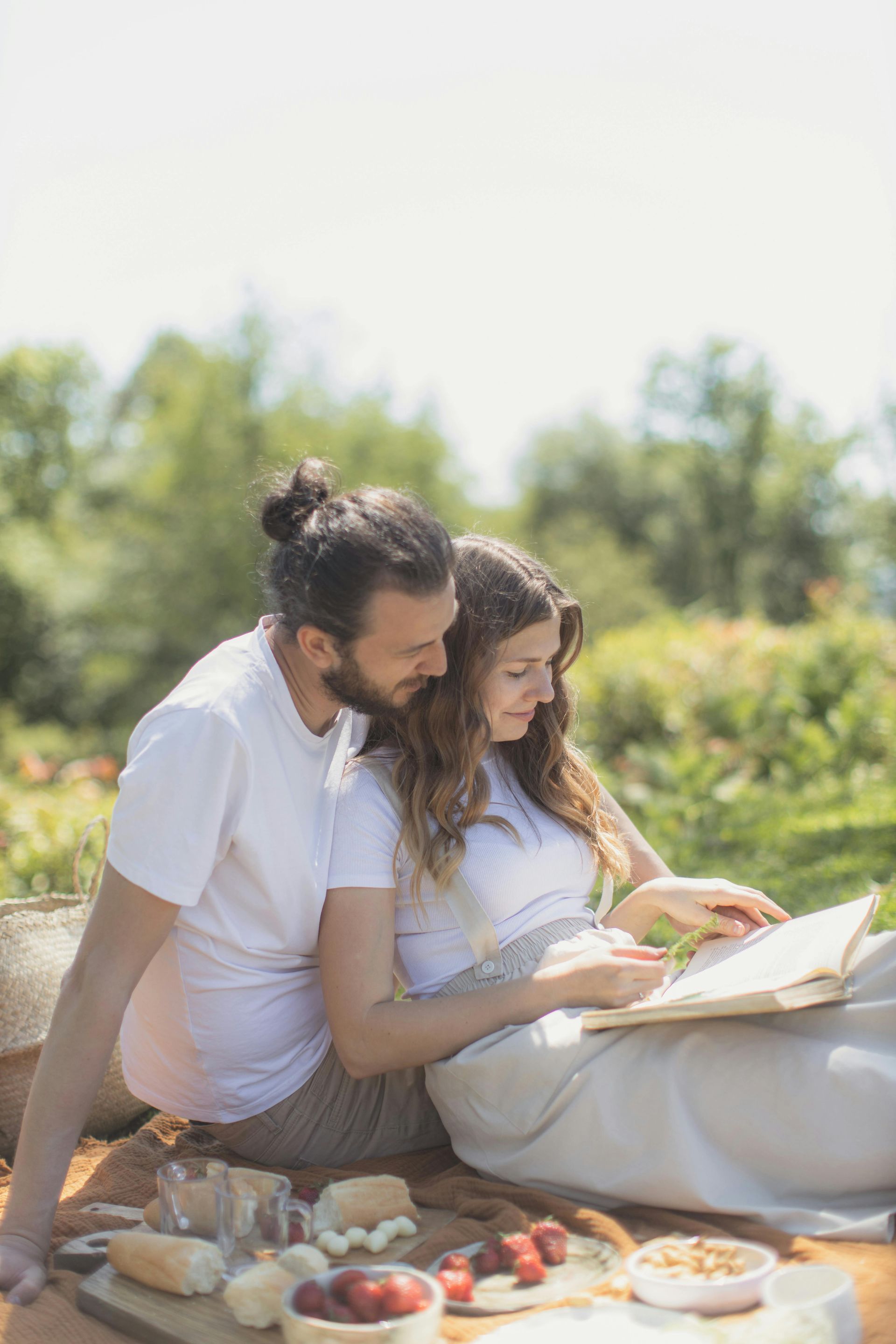 A couple sits on a picnic blanket in a sunlit meadow, reading a book together surrounded by snacks.