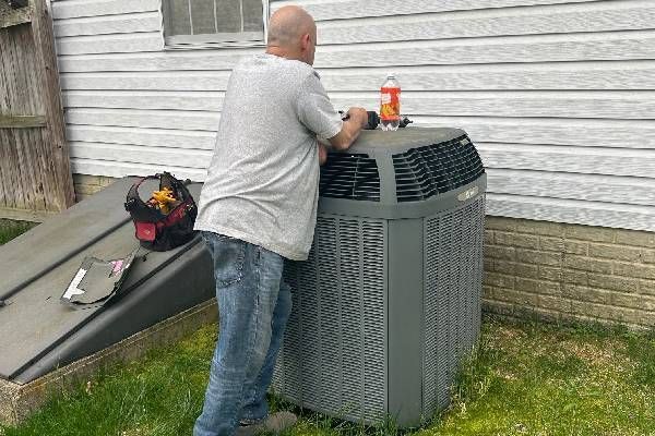 A person in a gray shirt and jeans leans over a gray outdoor HVAC unit next to a house, with a tool bag nearby.