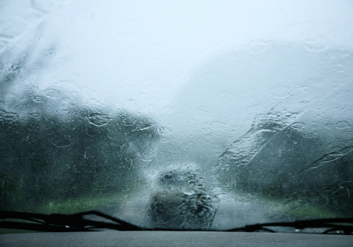 Rain-streaked windshield with blurred cars and trees in heavy downpour