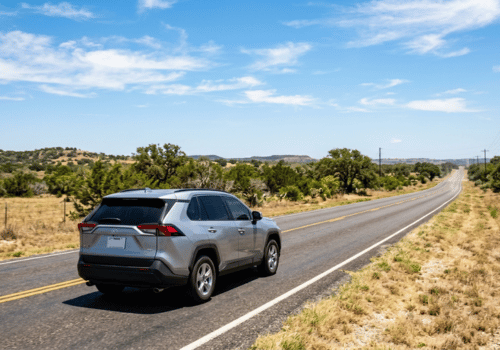 Silver SUV driving along a scenic rural highway through the Texas Hill Country on a clear summer day.