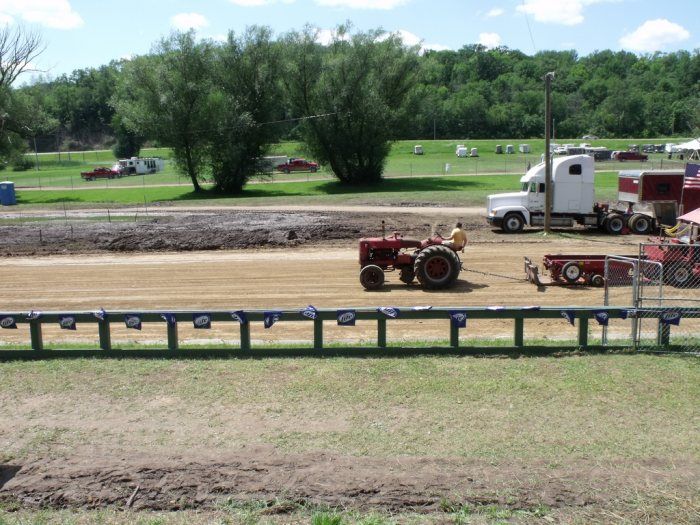 Red tractor pulling a weight sled at a tractor pull event. A semi-truck and trees are in the background.