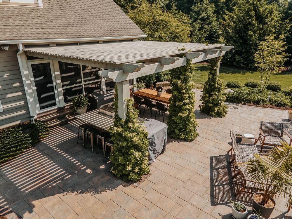 Patio with a pergola, dining table, and seating. The scene is outdoors with landscaping and a house in the background.