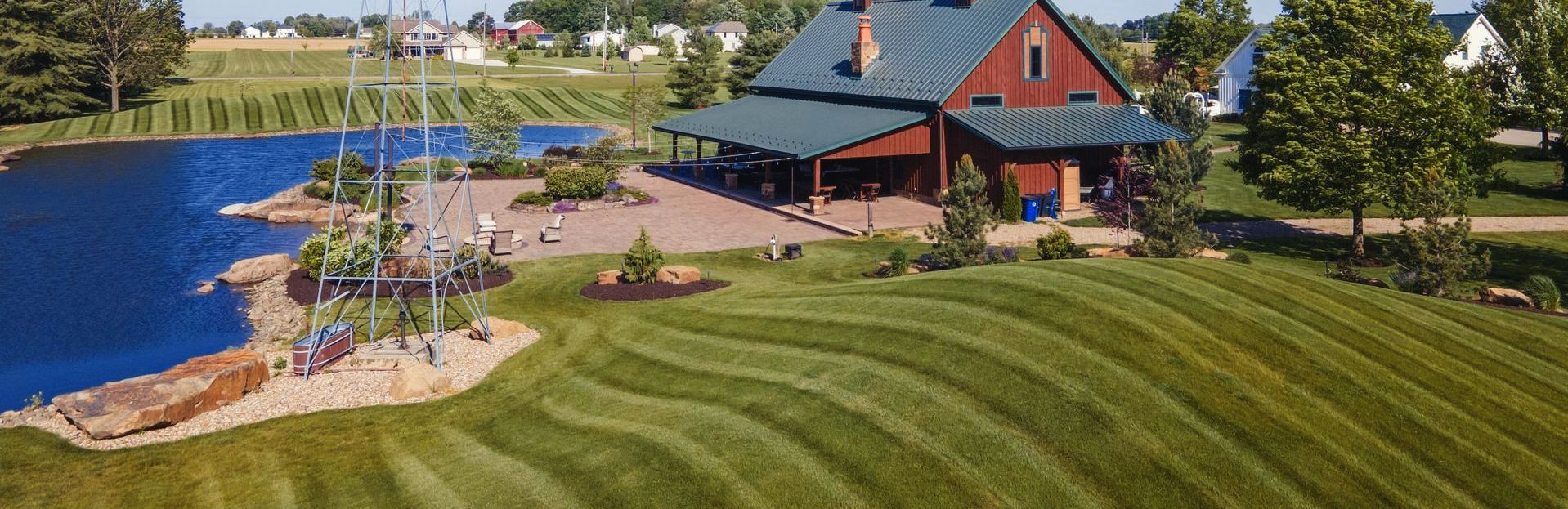 A red barn with a green roof is on a large green lawn near a lake.