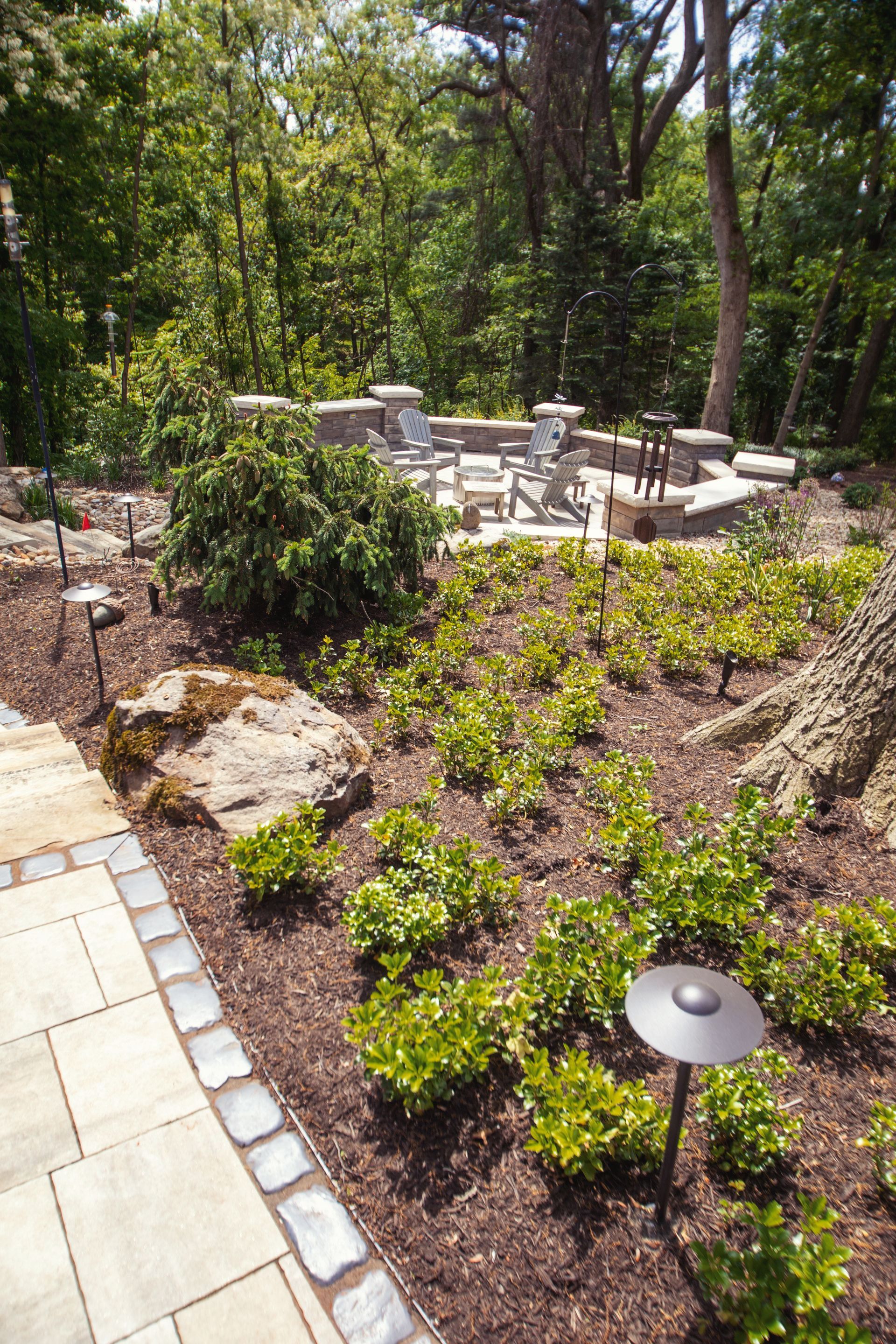 Landscaped garden with a stone patio, shrubs, and an outdoor seating area, with a forest backdrop.