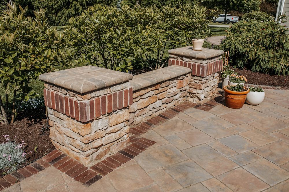 Stone and brick decorative garden wall with two pillars and built-in bench, with potted plants in a sunny outdoor setting.