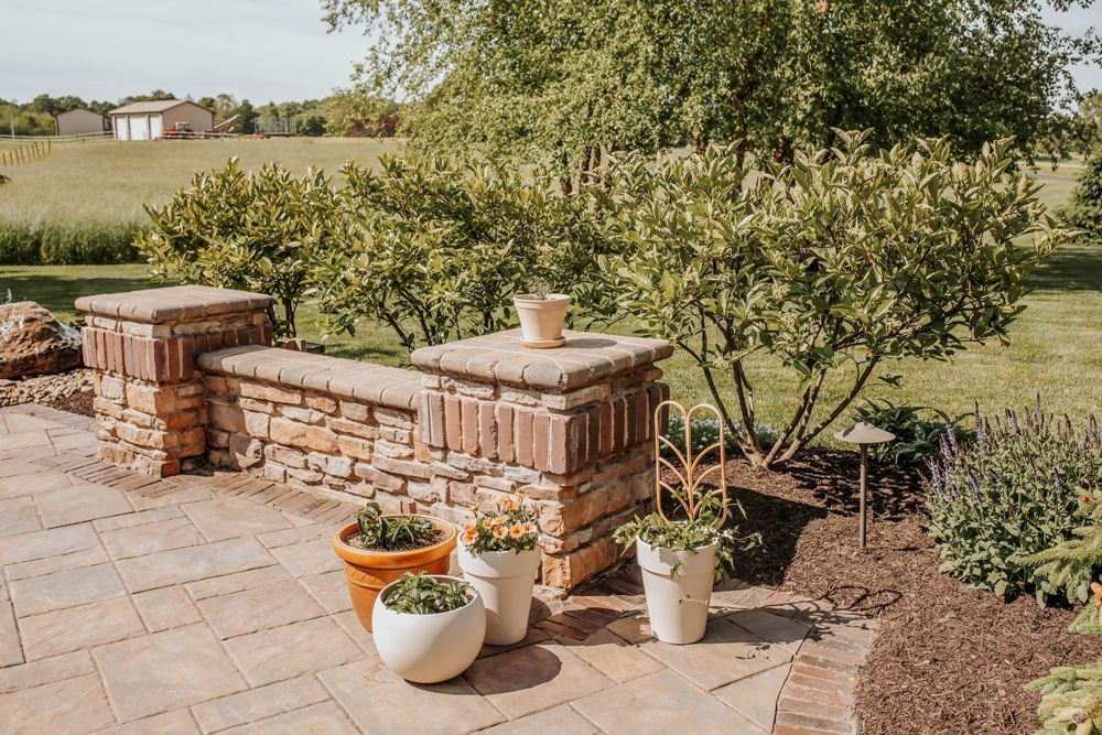 A brick patio with potted plants, a stone wall, and a tree. In the distance, there is a field and buildings.