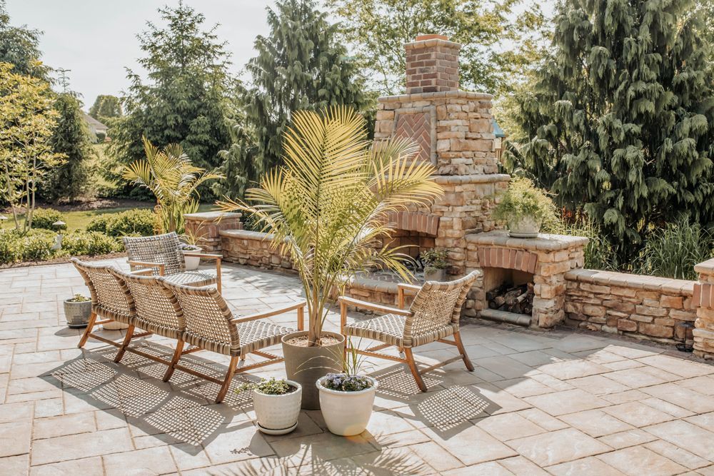 Patio with stone fireplace, potted plants, and a brick floor, set in a sunny outdoor area with trees in the background.