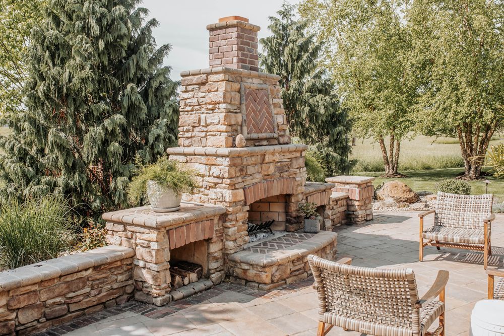 Outdoor stone fireplace and patio with seating, surrounded by trees and greenery.