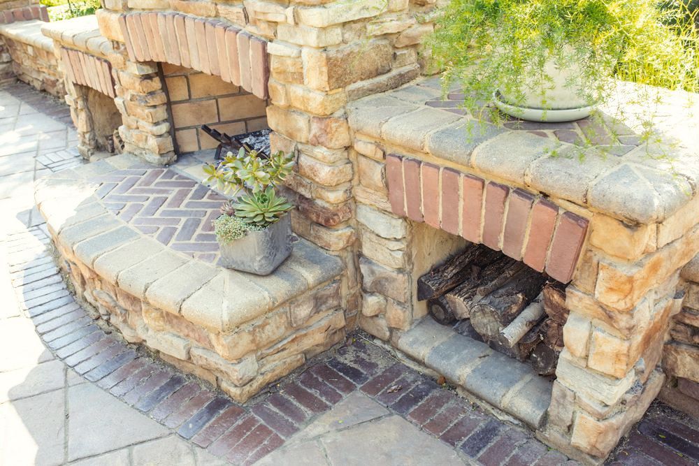 Outdoor brick fireplace and grill area. Stacked stone base with a brick arch over the fireplace opening, holding firewood.