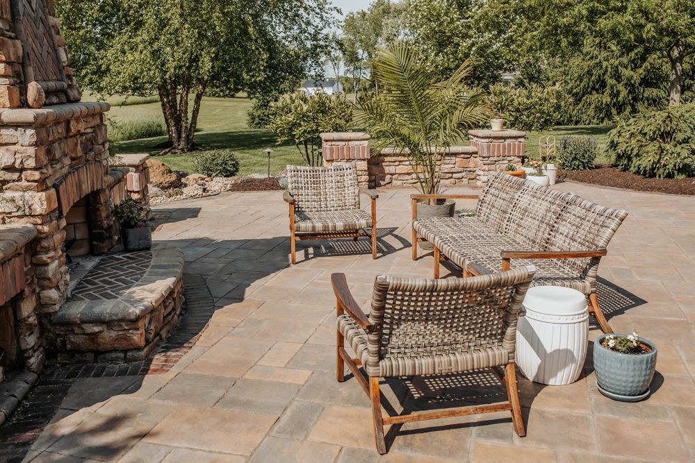 A patio with stone fireplace, seating area with wooden chairs and sofa, surrounded by trees and greenery.