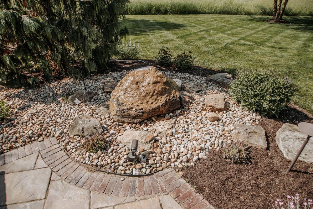 A garden bed with a large rock feature, surrounded by gravel, mulch, and paving stones, with green grass and bushes.