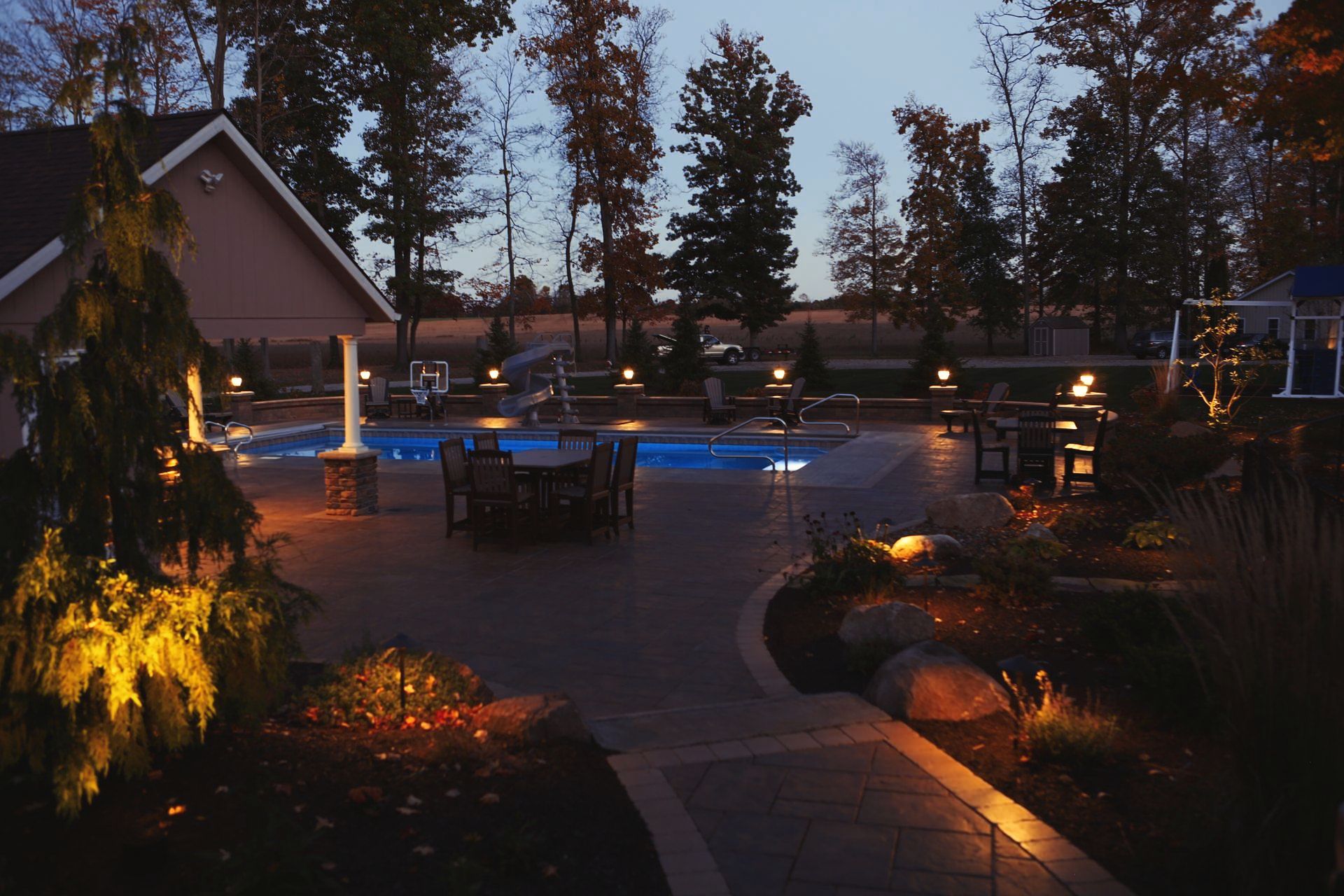 Night view of an outdoor patio with a pool. Warm lighting illuminates trees, seating, and landscaping.