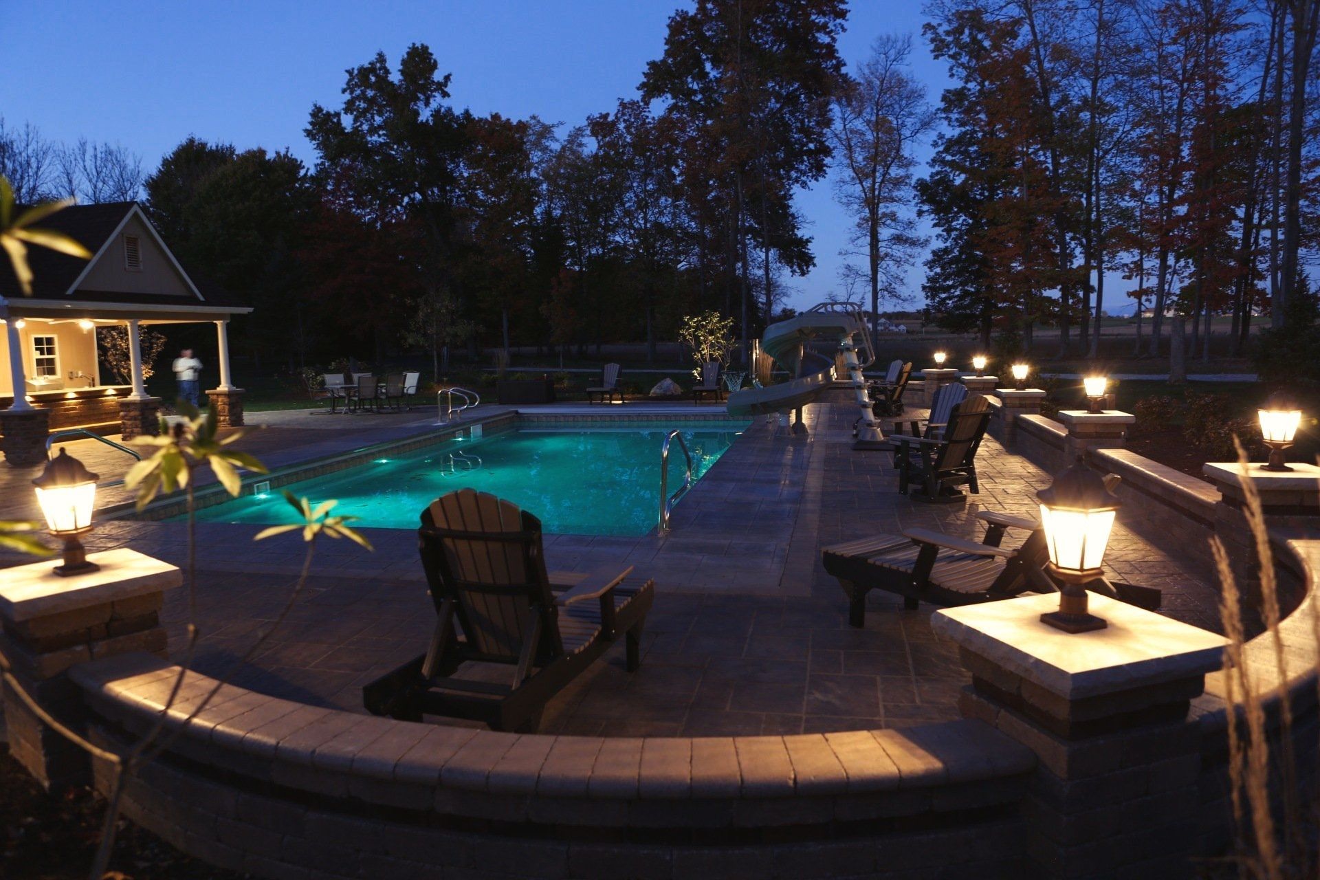 Night view of a pool area with a lit pool, seating, and decorative lights around the perimeter; trees in the background.