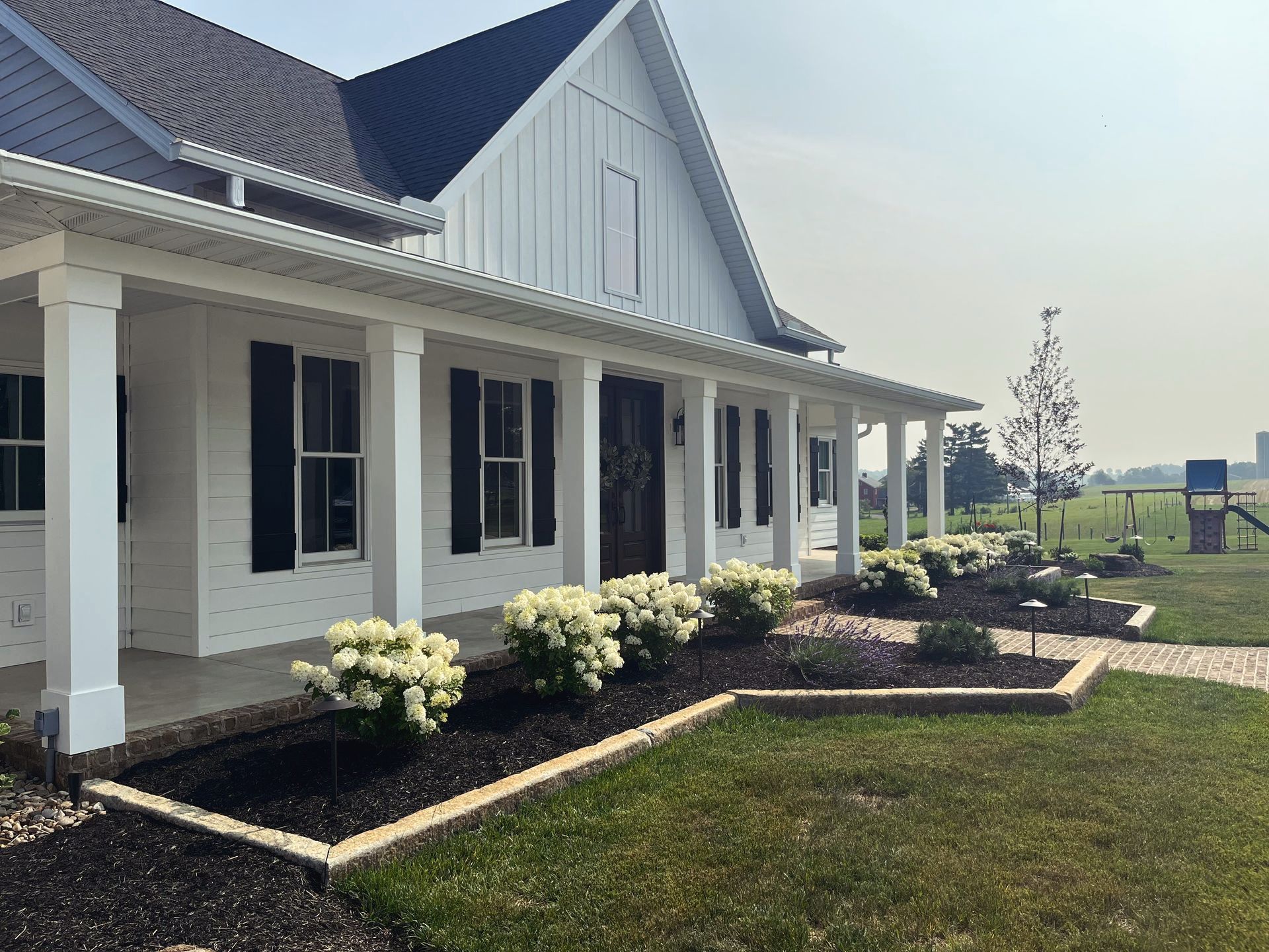White farmhouse with black shutters, a long porch, and a well-manicured lawn with flower beds.