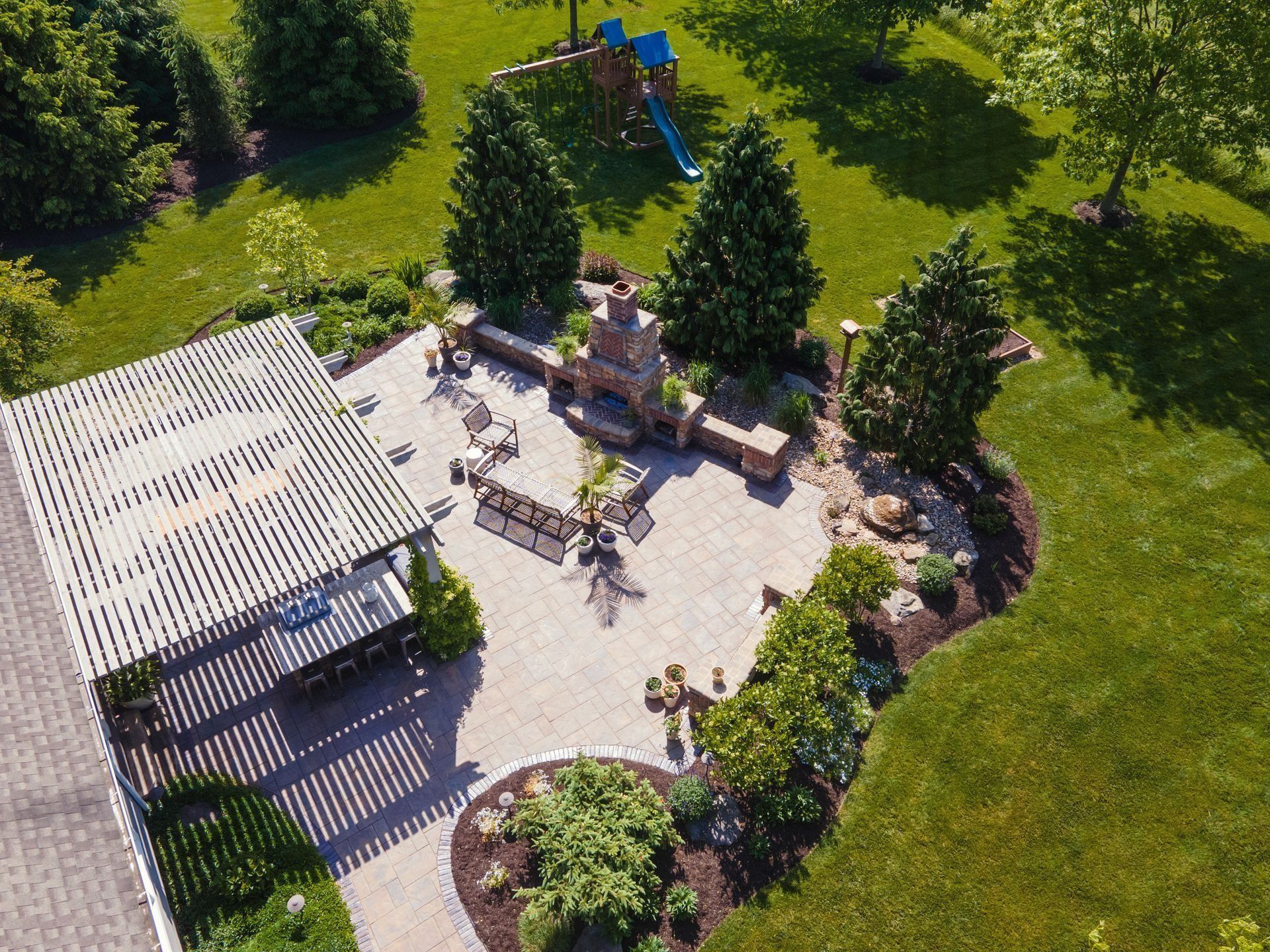 Overhead view of a backyard patio with stone pavers, an outdoor fireplace, pergola, and a small playset on a grassy lawn.
