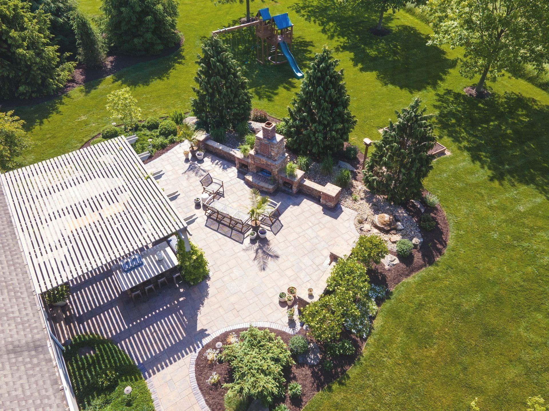 Overhead view of a backyard patio with pergola, fireplace, seating, and landscaping on a green lawn.