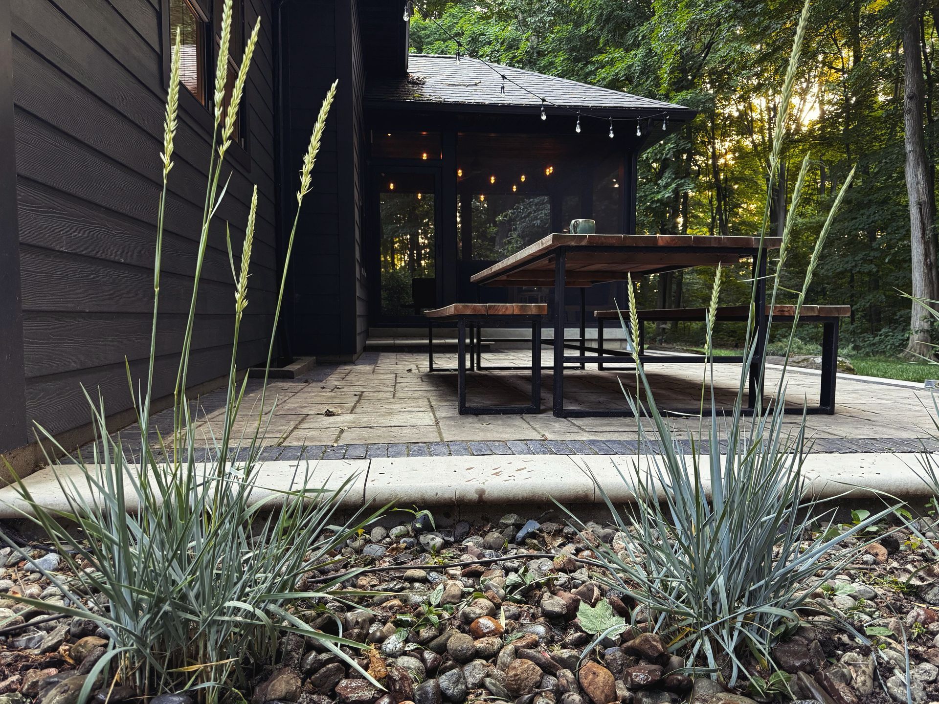 A dark-colored outdoor patio with a wooden table, surrounded by greenery and a small gazebo with string lights.