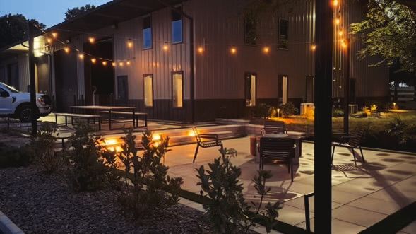 Night scene: outdoor patio with fire pits, seating, and a barn-like building strung with lights.